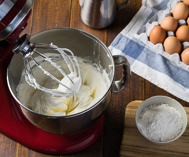 Kjøkkenmaskin som har vispet krem, egg og skål med mel står ved rundt maskinen på et bord.Foto