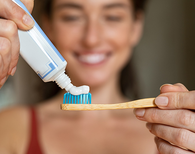 Tannkrem som blir presset ut av tannkremtube på tannkost med smilende dame i bakgrunnen.Foto