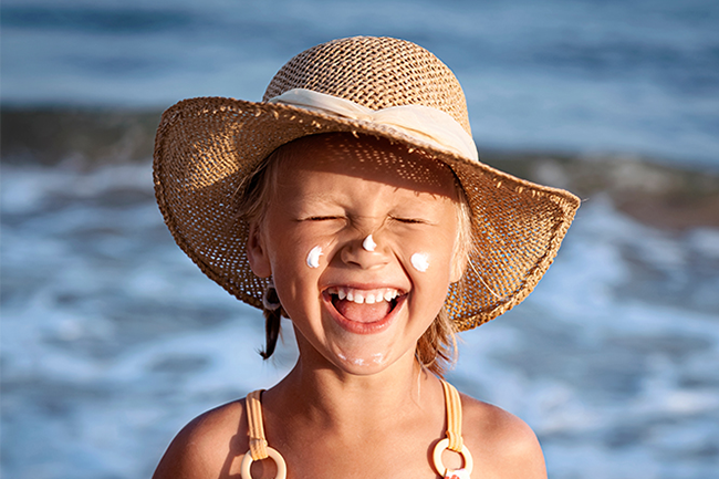 Glad jente med solhatt og to klatter med solkrem i kinnene foran et vann.foto